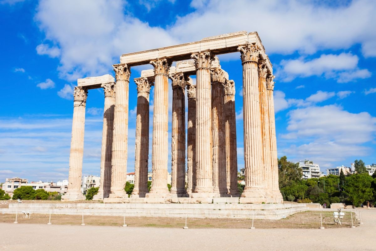 The image shows the Temple of Olympian Zeus in Athens, featuring several tall, ancient columns under a bright blue sky with scattered clouds.